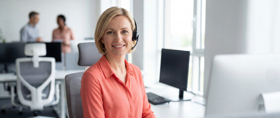 Fototapeta premium Female customer support agent in office wearing a bright coral shirt and black headset, smiling confidently at a modern desk for professional corporate business communication