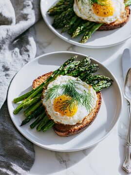 Overhead shot of two white ceramic plates featuring toasted bread topped with fried egg and fresh asparagus garnished with dill on a marble surface