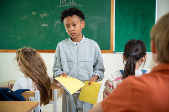 African American schoolboy handing out yellow notebooks to his classmates in classroom. Helpful multiethnic student distributing study materials or books during an elementary lesson.