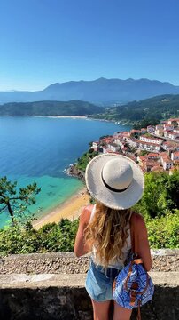 Woman tourist with straw hat and backpack looking at scenic aerial view of lastres village in asturias spain