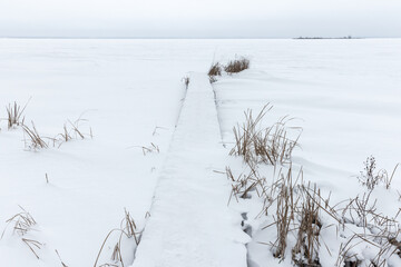 Naklejka premium A narrow snow-covered boardwalk leads across a vast frozen lake edged with dry reeds, evoking stillness, solitude and minimal winter beauty under a pale, overcast sky