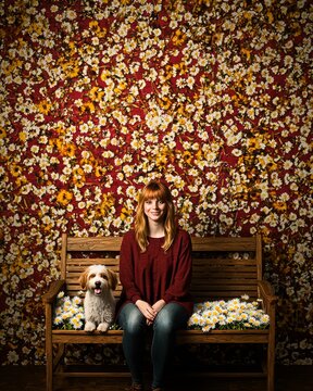 Young Woman with Red Hair Sitting on Wooden Bench Beside Sheepdog Against Floral Background
