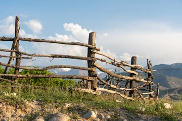 An old, weathered wooden fence made of rough tree branches and logs stands on a grassy, rocky hill. © Dina Luk
