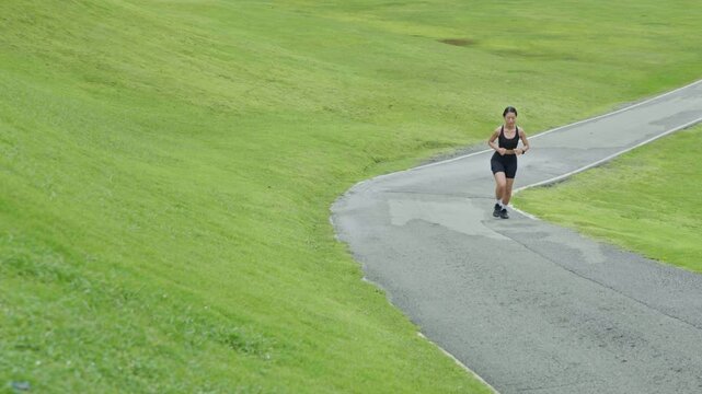 Woman running in the distance along a winding path in a green outdoor landscape, representing fitness, healthy lifestyle, endurance and longevity.