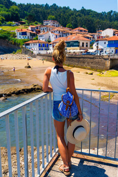 Woman tourist with backpack and straw hat looking at scenic beach and coastal village in Asturias, tour tourism, trave, destination in Spain