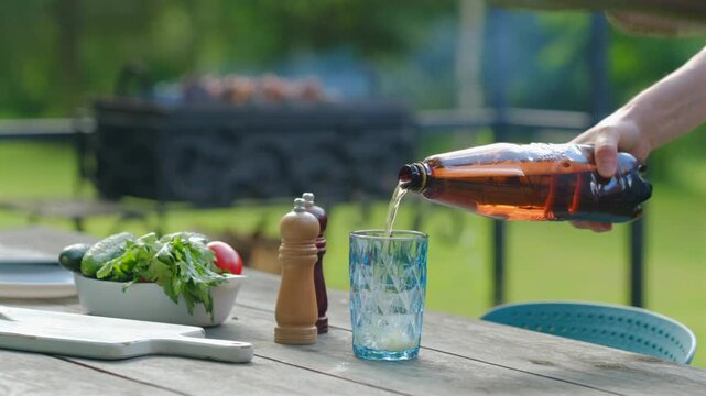 Pouring drink into glass at outdoor table during sunny day with fresh salad and grill in background