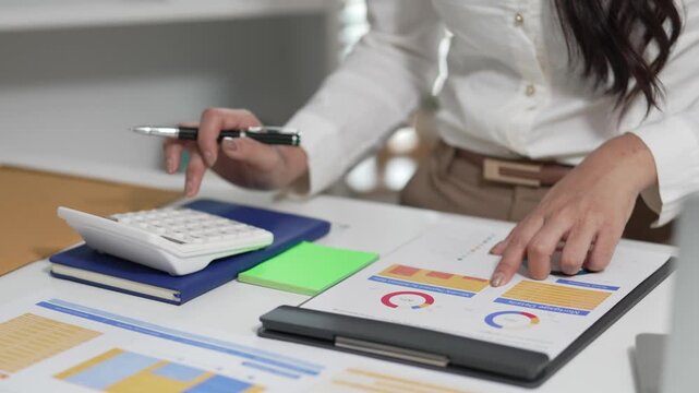 A smiling Asian businesswoman is working with a laptop and financial documents at her desk in the office.