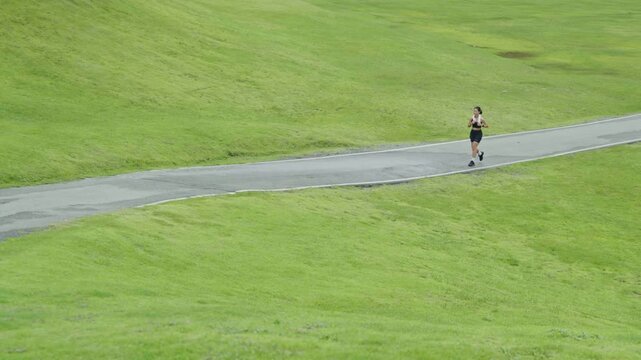 Woman running in the distance along a winding path in a green outdoor landscape, representing fitness, healthy lifestyle, endurance and longevity.