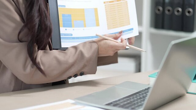 A smiling Asian businesswoman is working with a laptop and financial documents at her desk in the office.