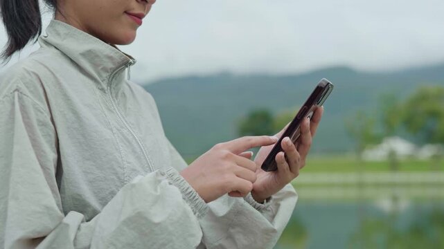 Close-up of a woman using a smartphone outdoors near a reservoir, representing healthy lifestyle, fitness tracking, active living and longevity.