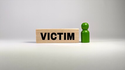 A wooden block printed with the word "VICTIM" stands next to a single green pawn figure against a plain white background, symbolizing human vulnerability. © FaizAhmad