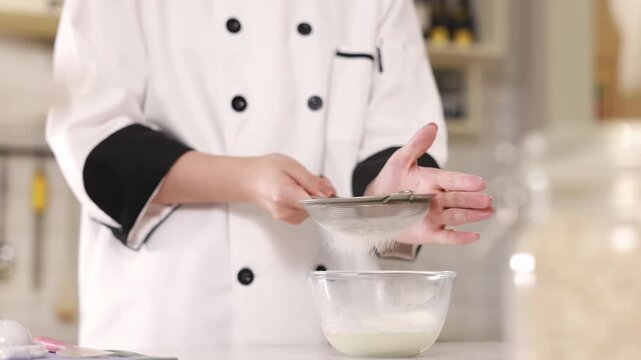 Pastry chef baker sift flour through a metal sieve into mixing white glass bowl to aerate for sponge cake batter, preparing dough for cooking, baking culinary preparation recipes food blogs in kitchen