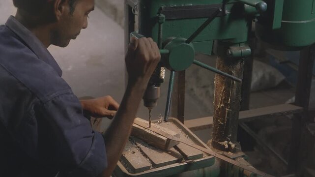 A close up indoor shot of an Indian Asian male worker or craftsman drilling a hole in a metal rod or grille with precision using bench mounted drill press machine in a fabrication workshop. 