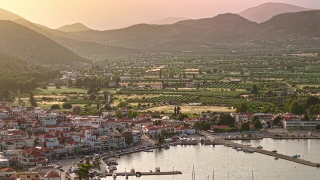 Aerial view of Ermioni port town, Greece at sunset. Cars move along the waterfront harbor while agricultural fields stretch toward mountain hills