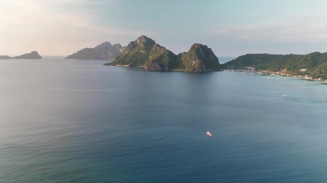 Beautiful high-angle drone shot of traditional paraw sailboats with striped sails cruising on calm sea water near dramatic limestone mountain islands under a soft sunset sky in the Philippines.