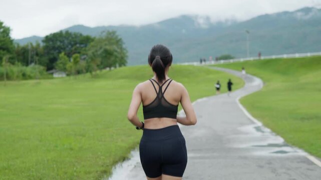 Back view of a female runner jogging along a winding park road with green hills and mountain scenery, representing outdoor fitness, endurance and healthy lifestyle.