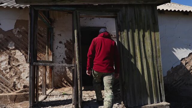 A man enters a lonely abandoned and dilapidated house on an abandoned farm to explore it, a gentle breeze rustling the branches of the bushes, early spring, cool sunny day