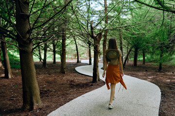 Naklejka premium Woman walking on winding path through forest with backpack and hat, trees overhead and sunlight filtering, nature trail scene for travel, solitude and summer outdoor exploration.