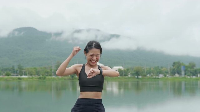 Female runner celebrating while checking her smartwatch after outdoor exercise by a lake, representing fitness achievement, health tracking, wellness and longevity.