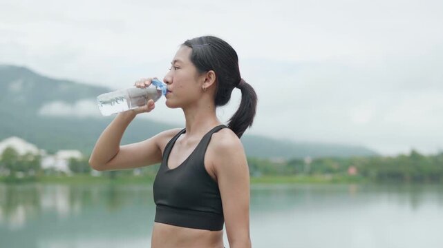 Female runner drinking water after outdoor exercise near a lake, representing hydration, fitness recovery, healthy lifestyle, wellness and active living.