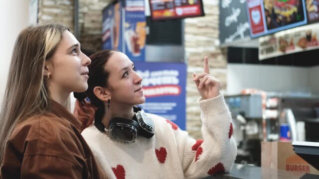 Two young women choose food from a menu on a self-service kiosk screen in a fast food restaurant.