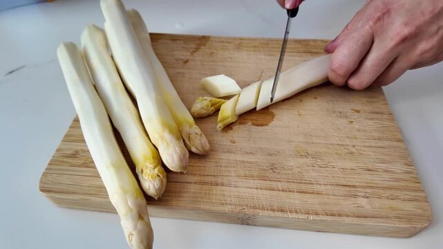 Cutting white asparagus into pieces on board. Slicing fresh asparagus stalks with a knife.