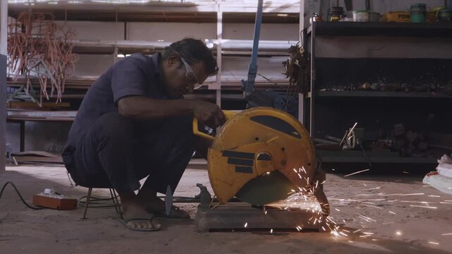 A wide indoor shot of a male Indian Asian worker using a chop saw to cut a steel rod to size, bright sparks fly by as abrasive wheels slice through the metal rod in a fabrication workshop.