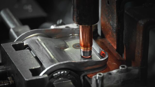 Close-up spot welding electrode contacting stamped steel automotive bracket in heavy fixture, showing heat tinted copper tip, machined clamp detail, and industrial assembly setup