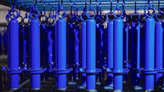 Rows of cobalt blue cylindrical metal parts hanging on overhead conveyor inside automotive components plant for powder coating finishing and industrial surface treatment