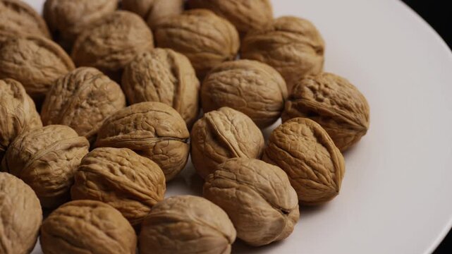 A high angle close up shot showing many whole brown walnuts arranged on a clean white ceramic plate