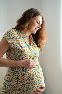Pregnant woman holds her abdomen with concerned, pensive expression indoors. Patient feeling her belly shows worried vulnerability and medical concern about health challenges at forty in hospital