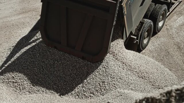 Rear-left elevated view of heavy-duty dump truck unloading crushed stone into growing gravel mound at production site, tailgate open, bed tilted, bright sunlight, dust rising