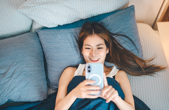 Happy young adult asian woman waking up in bedroom on morning enjoy checking smartphone social media Relax rest and leisure lifestyle