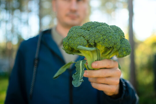 A man holds fresh broccoli in front of his face in a closeup outdoors, presenting sustainable eating. The consumer shows a healthy, vibrant vegetable in natural daylight with greenery behind.