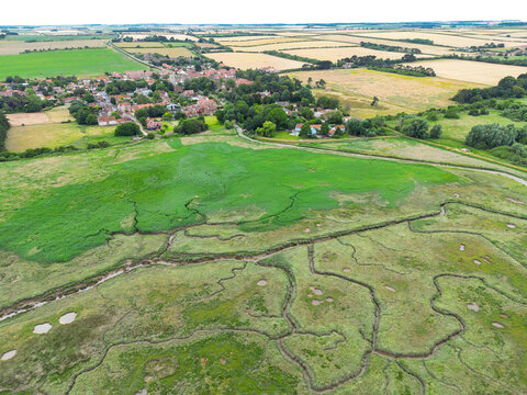 Drone view of large areas of salt marshes and salt water tributaries seen on a flood plain on the north Norfolk, UK coastline. In the distance is the village of Brancaster.