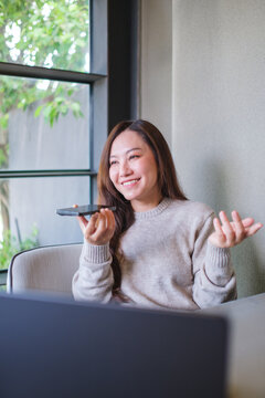 Vertical of a woman holding smart phone and using voice messaging, talking with ai assistant for Artificial intelligence concept while working on laptop