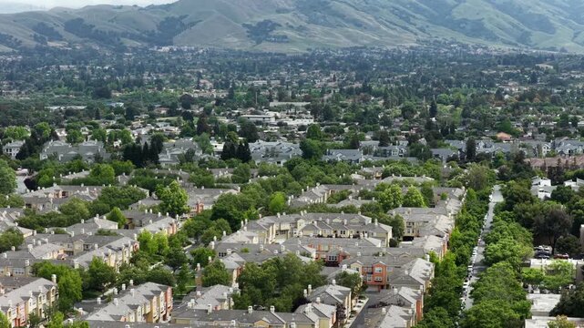 Aerial drone view over modern residential neighborhood in Fremont, California with rows of townhouses, tree-lined streets and dense suburban development on a sunny day. Wide shot. Quiet scene.