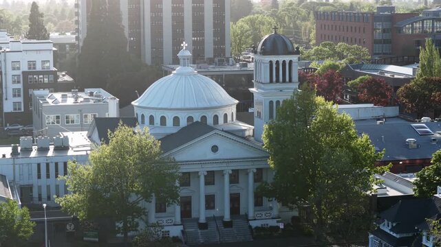 Historic domed church with white columns anchors downtown Eugene, Oregon at golden hour, surrounded by leafy green trees plus mid-rise buildings under warm evening light. Aerial drone wide shot