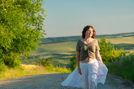 Happy young woman in white skirt walking on countryside road at sunset