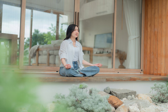 Young Asian woman practicing mindfulness meditation with closed eyes on home terrace beside rock garden, calm morning wellness routine.