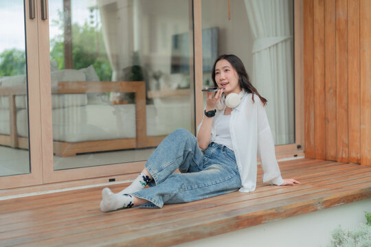 Young Asian woman speaking into mobile phone on speakerphone with headphones around neck, communicating outdoors on house terrace.
