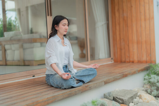 Young Asian woman taking quiet break to meditate on wooden terrace, mental health self care and emotional balance in daily life.