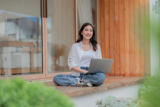 Young Asian digital nomad typing on computer outdoors, remote work from vacation home deck with green garden, flexible workplace.