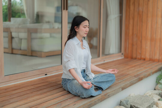 Side view of young Asian woman doing breathing exercise in cross legged pose on wooden deck, quiet moment of zen relaxation at home.