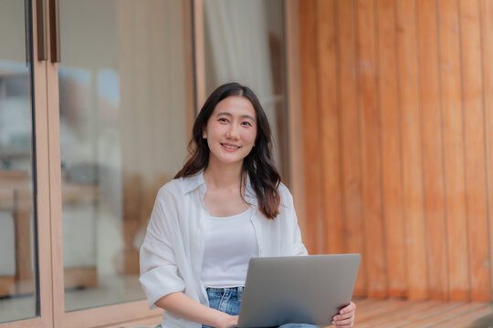 Cheerful young Asian freelancer looking at camera with computer on lap, self employed worker enjoying job outside modern home.
