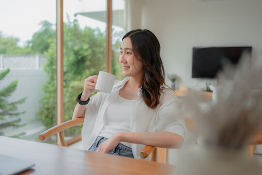 Young Asian woman enjoying quiet morning with hot drink, gazing through window at nature, slow living moment in modern interior.
