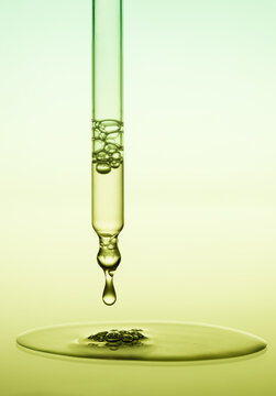 Dropper with a drop of cosmetic serum or gel falling and spread on green background.