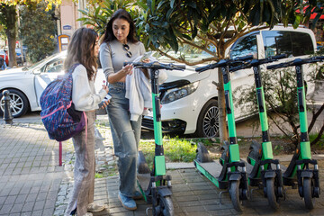 Two people discuss electric scooters on a city street with trees and cars in the background during the day © artmim