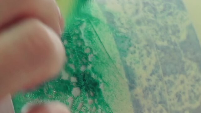 Close-up of a hand skillfully brushing vivid green pigment onto a delicate textured sheet. The creative process unfolds as gentle strokes bring a rich emerald design to life.