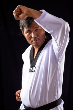 Taekwondo black belt demonstrates high block technique in white dobok against dark studio background, highlighting discipline, defense, and martial arts skill
